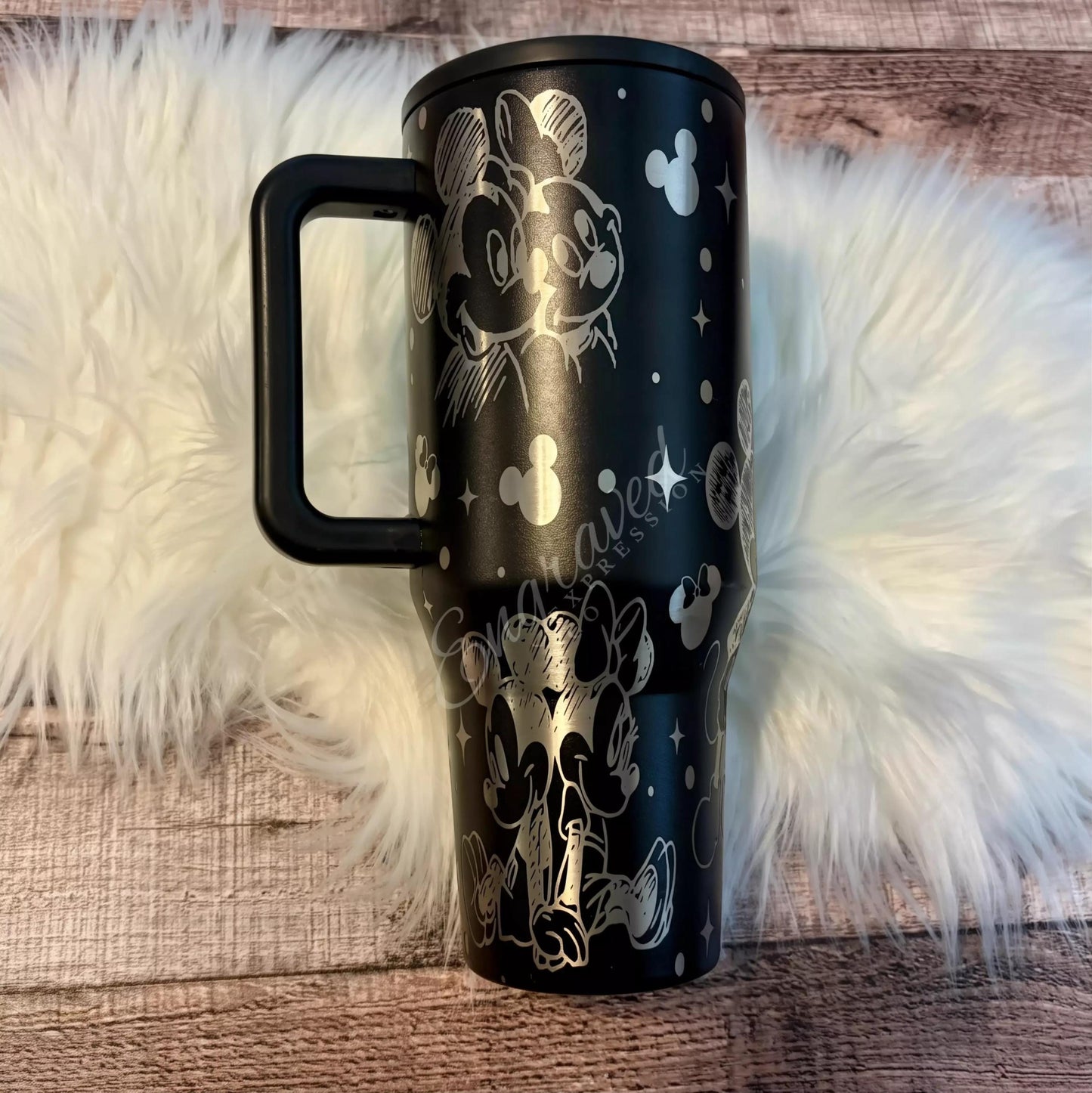 A black tumbler mug with a handle and a design featuring Mickey Mouse and Minnie Mouse characters on it, placed on a white fur rug.