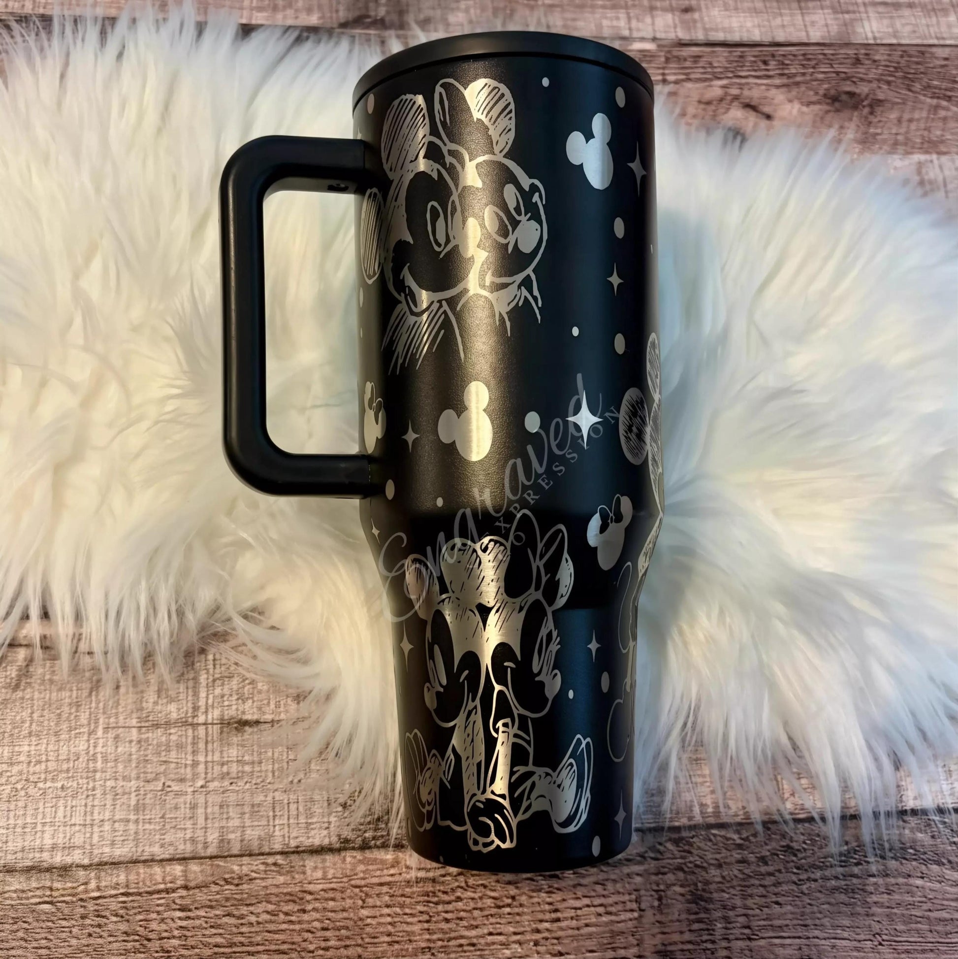 A black tumbler mug with a handle and a design featuring Mickey Mouse and Minnie Mouse characters on it, placed on a white fur rug.