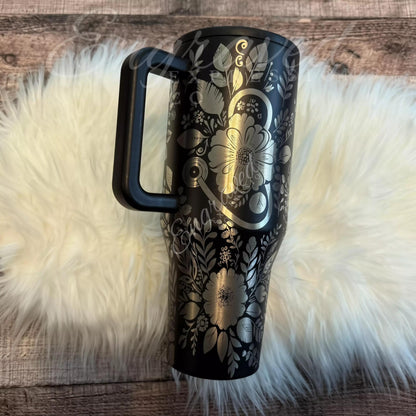 A black and silver floral patterned thermos mug with a black handle is placed on a white furry surface.