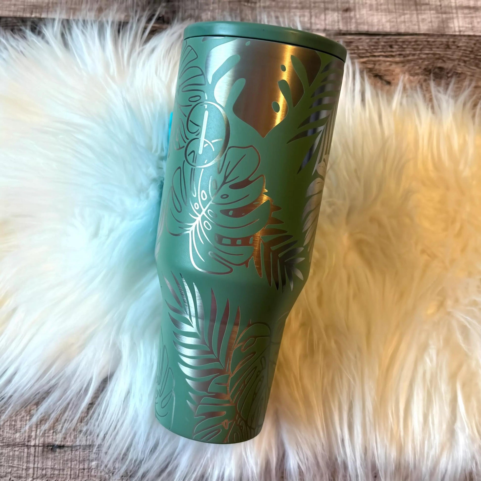 A green tumbler with a leafy palm tree design sits on a white furry surface.