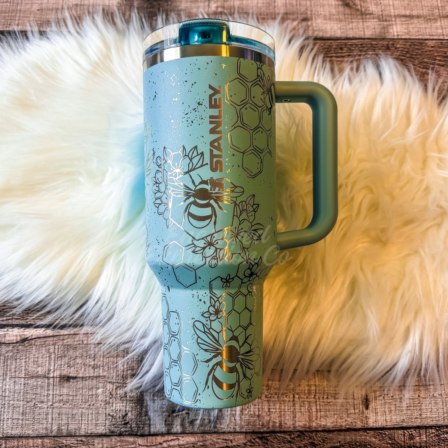 A blue Stanley tumbler mug with a handle and a clear lid, placed on a white furry surface.