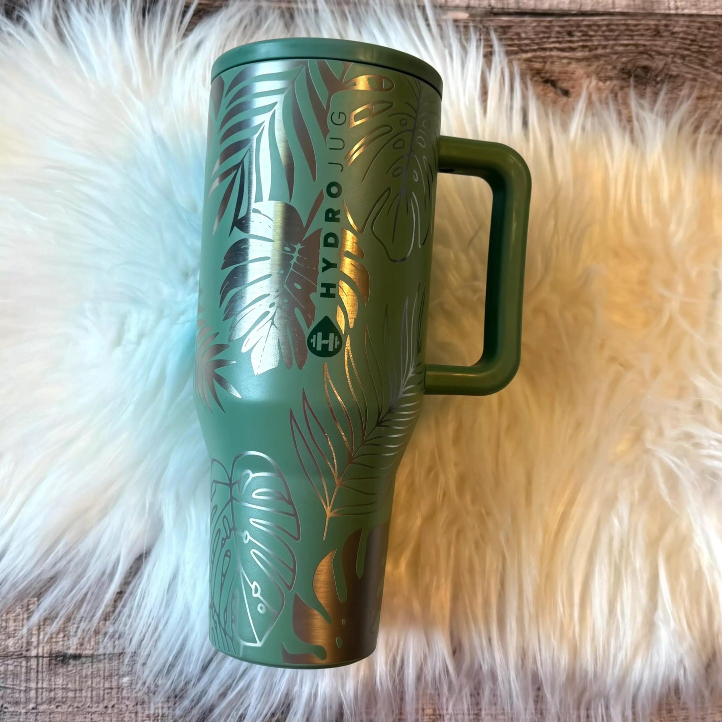 A green travel mug with a handle and a palm leaf design, placed on a white furry surface.