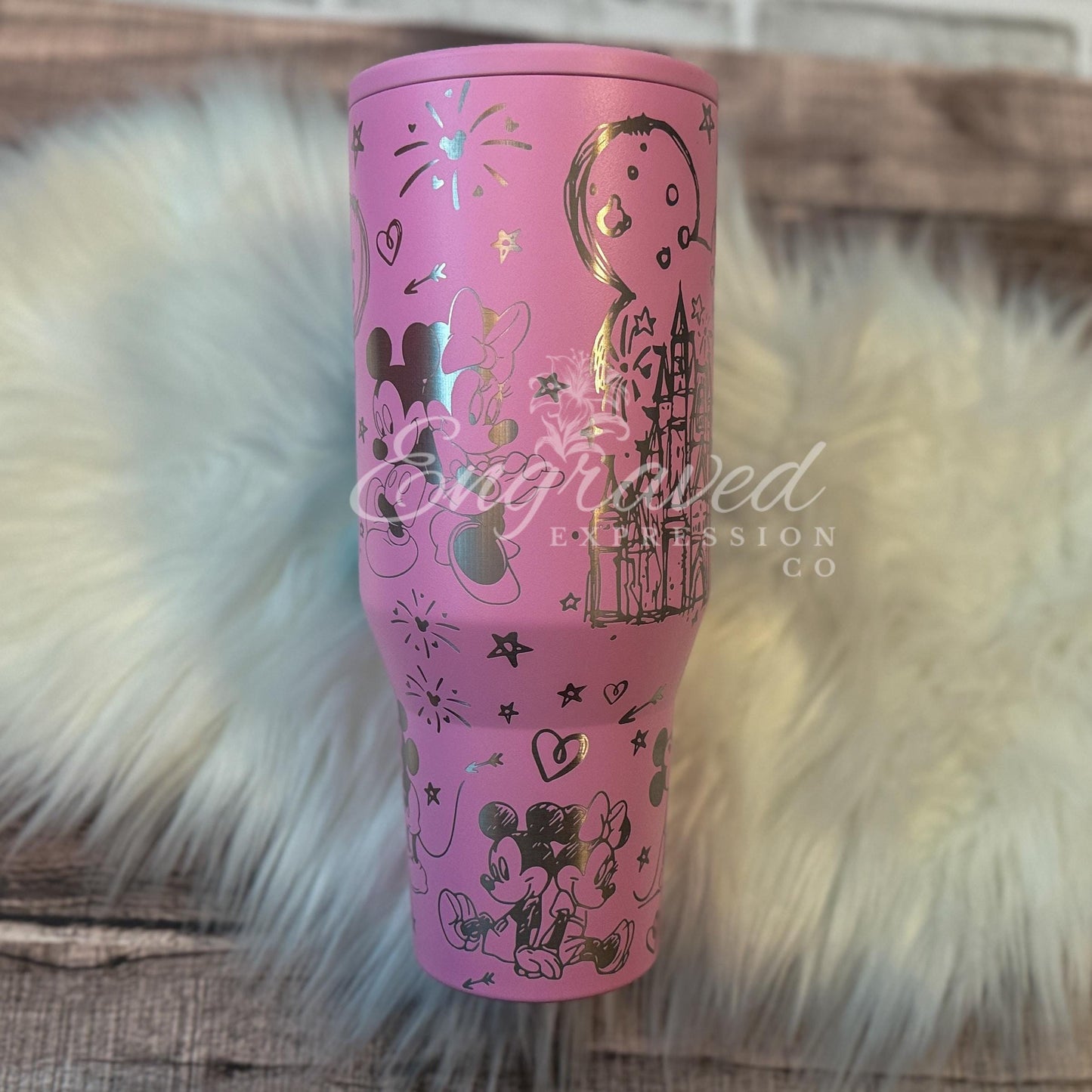 A pink tumbler with various Disney-themed designs and characters printed on it, placed on a white furry surface.