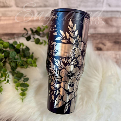 A black tumbler with a floral design on it, placed on a white fur surface next to some greenery.
