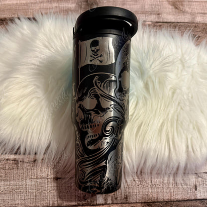 A black and silver tumbler mug with a skull and crossbones design, placed on a white furry surface.