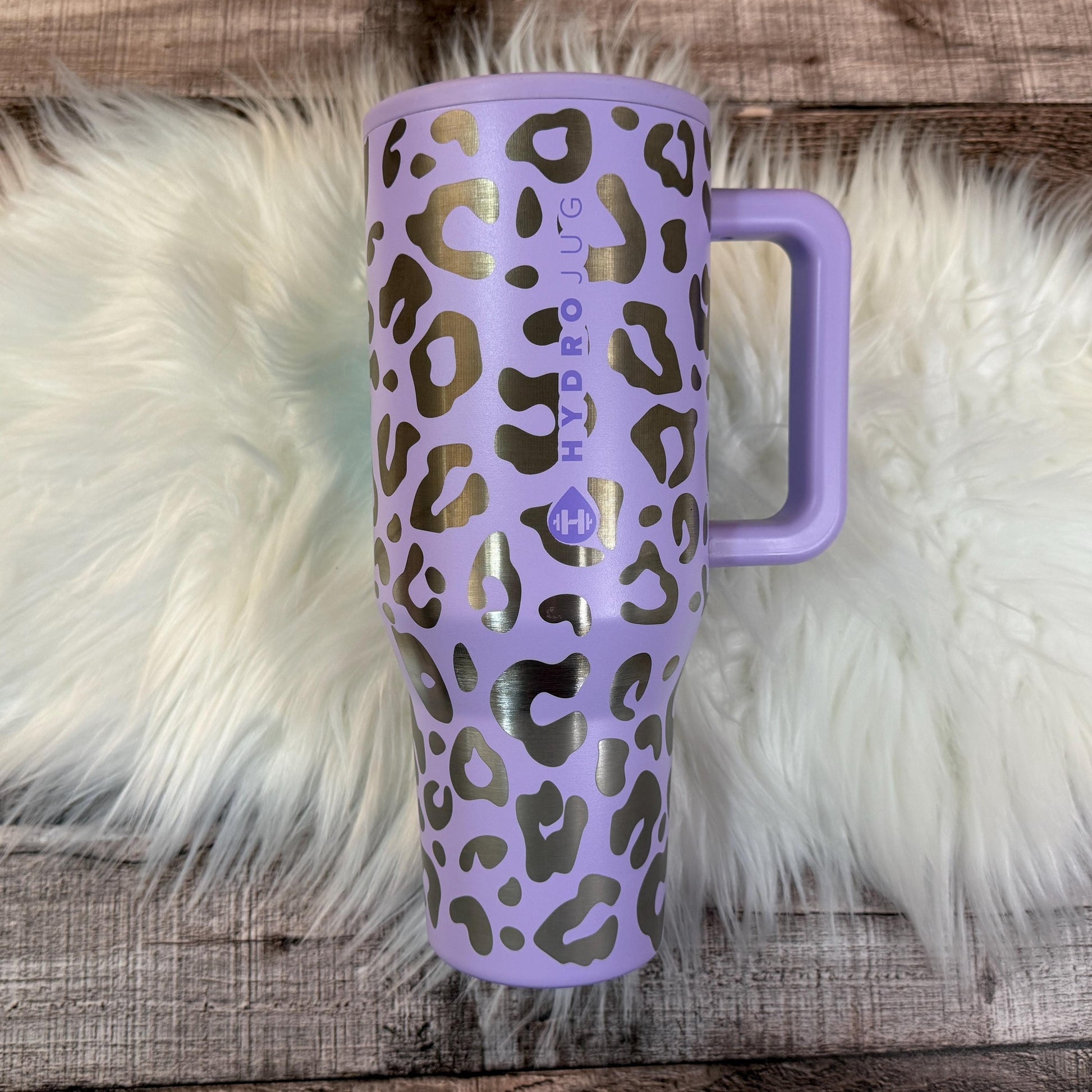 A purple and black leopard print thermos mug with the text "HYDRO JUG" printed on it, placed on a white fur rug.