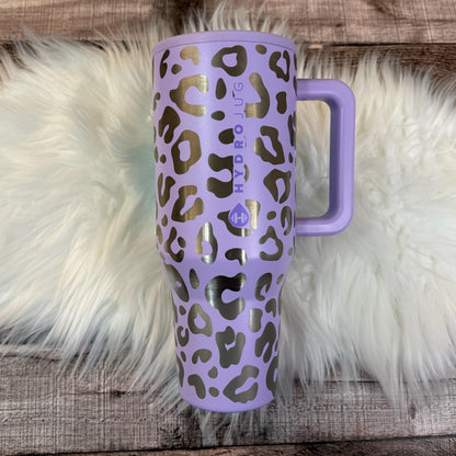 A purple and black leopard print thermos mug with the text "HYDRO JUG" printed on it, placed on a white fur rug.