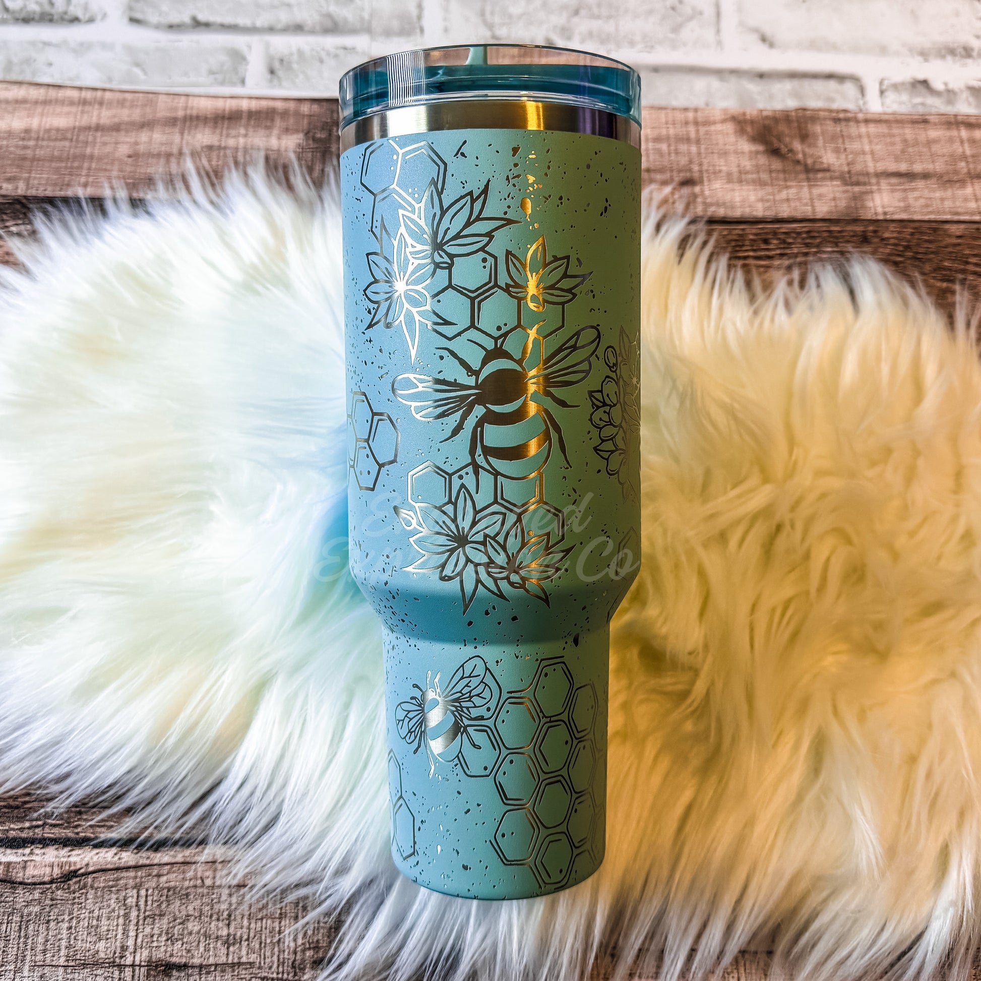 A turquoise insulated tumbler with a honeycomb design and a bee printed on it, placed on a fluffy white surface.